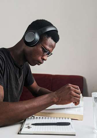 A man is sitting at a table with a notebook, pen, and glass of water, wearing headphones.