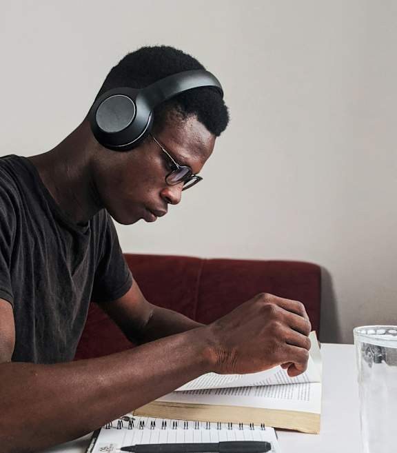 A man is sitting at a table with a notebook, pen, and glass of water, wearing headphones.
