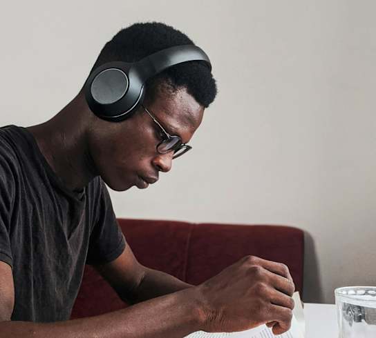 A man is sitting at a table with a notebook, pen, and glass of water, wearing headphones.