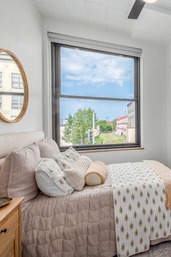 A bedroom with a bed, nightstand, and a window overlooking a street. at Life At Agora Apartments, Cleveland, Ohio