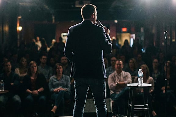A man in a dark suit is speaking into a microphone in front of a crowd in a dimly lit room at The Lofts at Elk Terminal Apartments, Buffalo