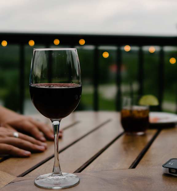 A glass of red wine is on a table with a person's hands in the background.