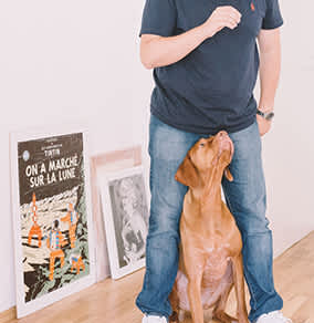 A man wearing a blue shirt and jeans holds a brown dog up in the air.
