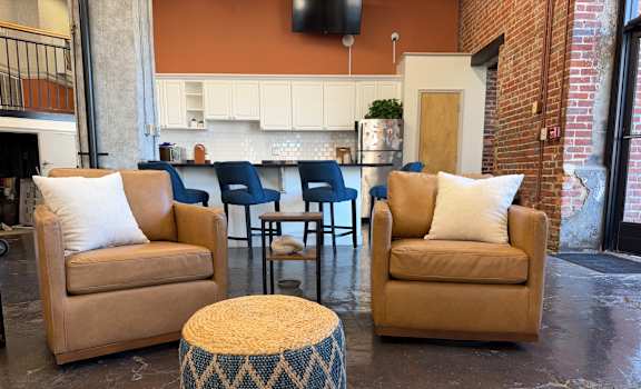 A living room with a tan couch, blue and white ottoman, and a brick wall.
