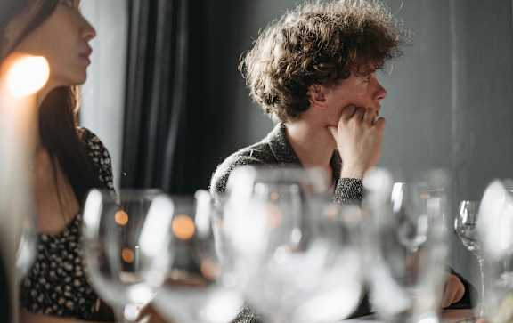 A man with curly hair is sitting at a table with wine glasses in front of him.