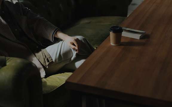 A person is sitting on a green couch with a coffee cup and a book on the table.