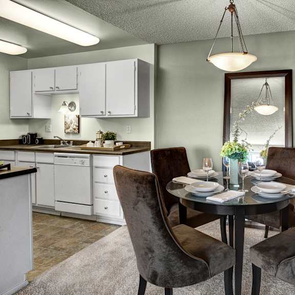 The dining room with plush dining chairs surrounding a dining table with fully set place settings and flowers, with the kitchen in the background at Overlook at Magnolia Apartments. 
