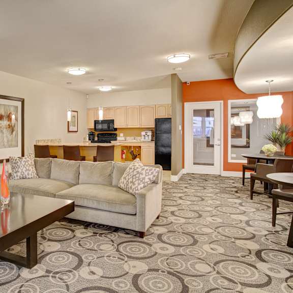 The resident lounge with a TV, plush couch, artwork and decorations, a coffee table, dining tables and chairs, and a community kitchen including spacious light cabinets, barstool chairs, and black appliances at Overlook at Magnolia Apartments. 