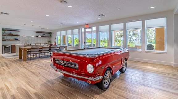 A red vintage car is parked in a spacious room with wood flooring and large windows.