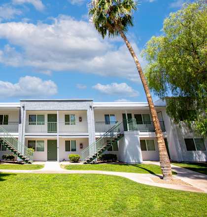 A white building with a balcony on the second floor is surrounded by green grass and trees.