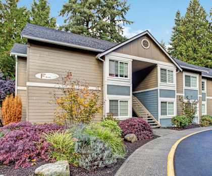 The beautiful exterior of the tan, gray, and white apartments and the front garden with flowers, trees, and shrubs with a sidewalk and road in front at Vermont Apartment Homes. at