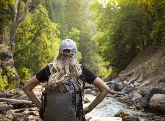 A woman with a backpack enjoys a hike by the river, surrounded by nature.