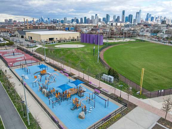 A playground with a blue surface and purple trim is surrounded by a fence and has a city skyline in the background.at 829 Garfield, New Jersey