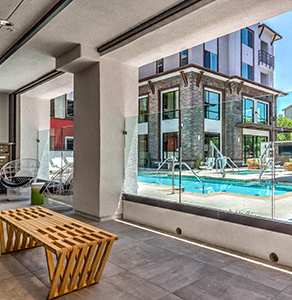 A wooden bench sits in front of a window with a view of a pool and a building.