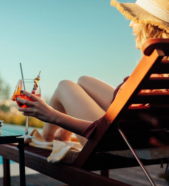 A woman in a straw hat is sitting on a chair with a glass of wine and a plate of food in front of her.