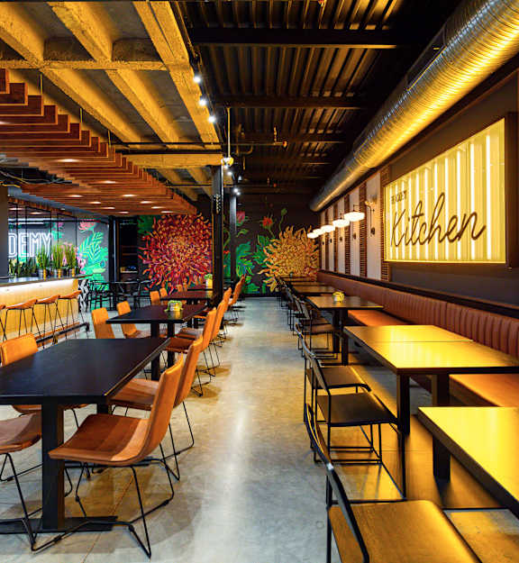 A restaurant with a long table and chairs with a neon sign that says kitchen.