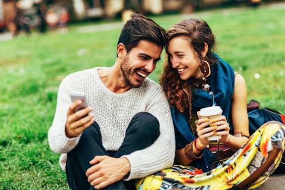 A man and a woman are sitting on the grass, each holding a cup, and the man is looking at his phone.