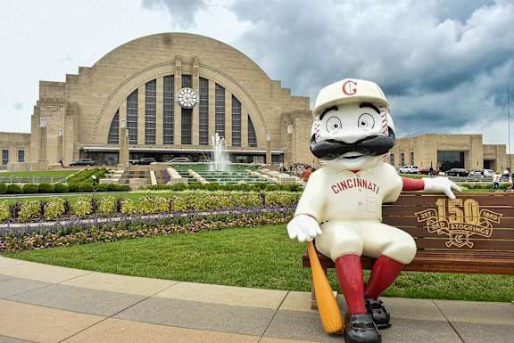 A mascot dressed in a Cincinnati baseball uniform is sitting in front of a building with a fountain.