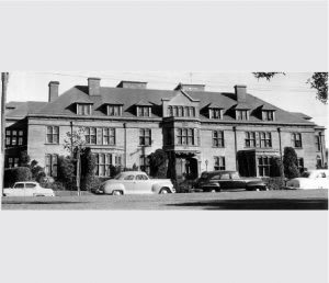 A black and white photo of a large house with a car parked in front.