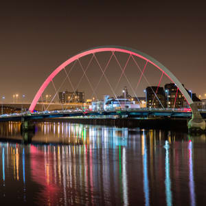 A bridge with a pink arch spans a body of water at night.