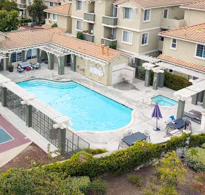 A tennis court is surrounded by a pool and apartment buildings.