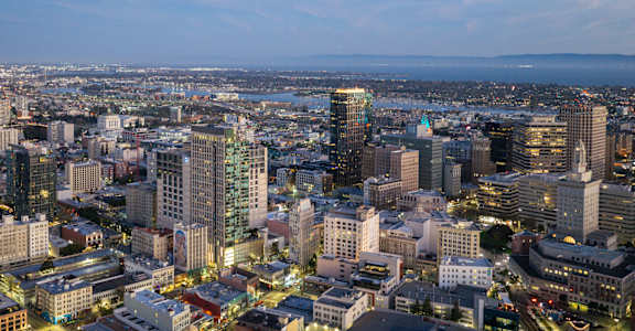 A cityscape at dusk with buildings illuminated and a body of water in the distance.