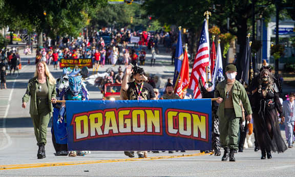 A group of people in military uniforms marching in a parade with a banner that reads "Dragon Con".