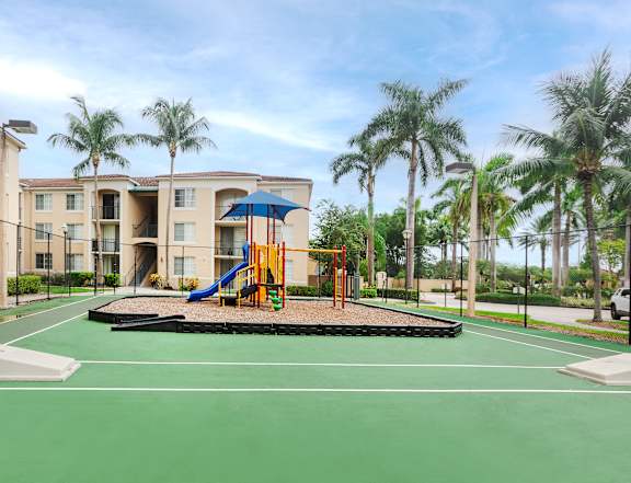 A playground with a slide and swings in front of a building with palm trees.