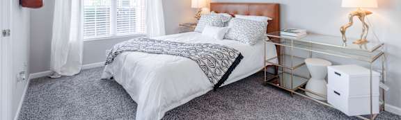 A bed with a white comforter and a black and white patterned blanket is in the foreground with a lamp and a window in the background.