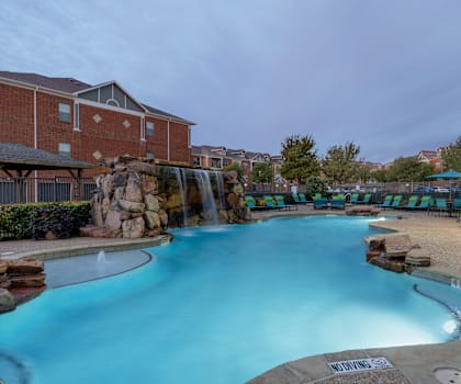 The large, turquoise outdoor pool with a large waterfall, green and blue pool chairs with greenery, trees, and the apartments in the background at the Landing at Mansfield Apartment Homes. 