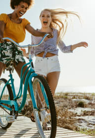 two women riding bikes on a boardwalk with the ocean in the background