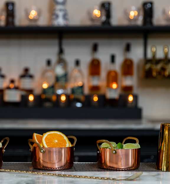 Three copper cups with garnishes sit on a bar counter in front of a shelf of bottles.