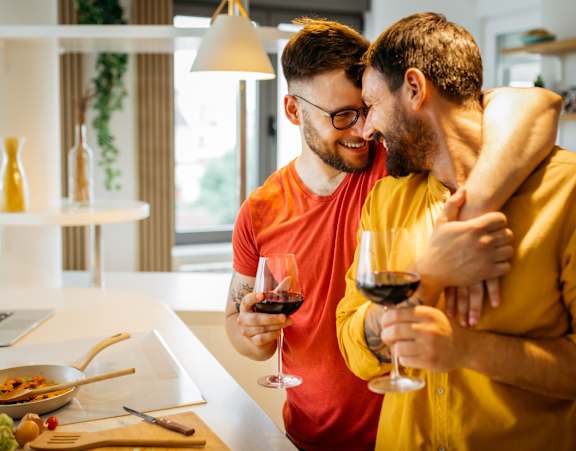 Two men are standing in a kitchen, one holding a glass of red wine and the other with his arm around the other man.