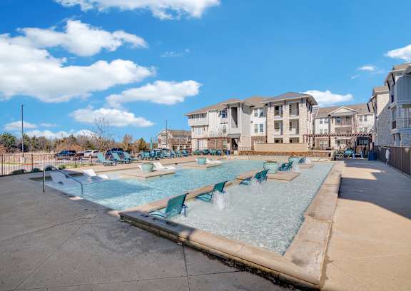 A swimming pool with chairs around it and apartment buildings in the background.
