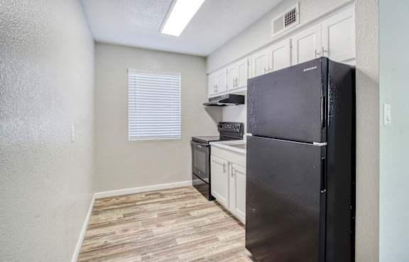A black refrigerator is in a kitchen with white cabinets and a window with blinds.
