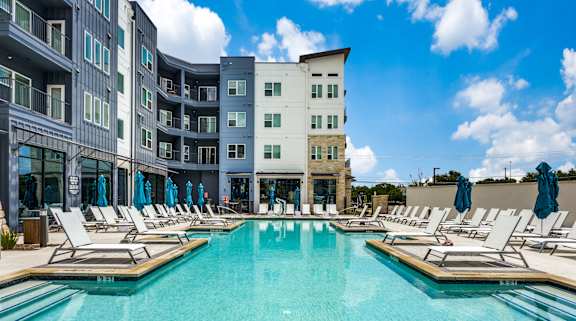 A large swimming pool with lounge chairs and umbrellas in front of a multi-story building.