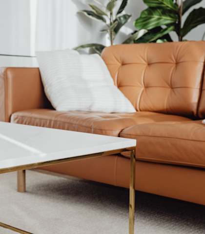 A white coffee table with gold legs sits in front of a brown leather couch with a white pillow on it.