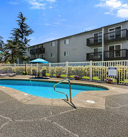 a swimming pool with tables and umbrellas in front of an apartment building at Pacific Park Apartment Homes, Edmonds, Washington 98026