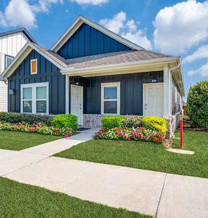 A house with a blue roof and white walls is surrounded by a well-manicured lawn and colorful flower beds.