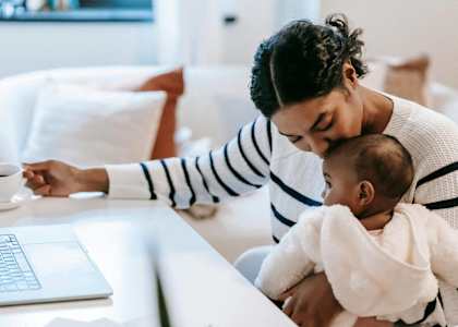 A woman holding a baby while sitting at a table with a laptop.