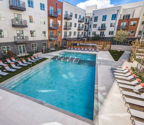 A swimming pool surrounded by sun loungers in front of apartment buildings.