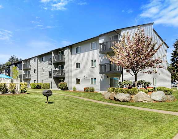 a grassy area with a tree in front of an apartment building at Pacific Park Apartment Homes, Edmonds, WA