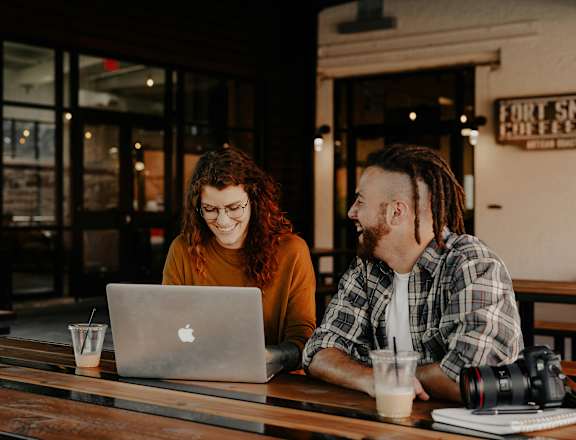 Two people sitting at a table with a laptop and a camera.
