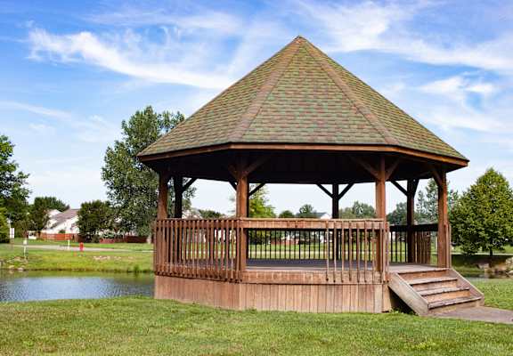 A gazebo with a brown roof and wooden pillars is situated in a grassy area with a pond in the foreground.