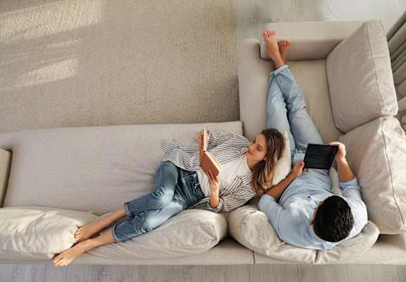 Two people lying on a couch, one holding a book and the other holding a tablet.