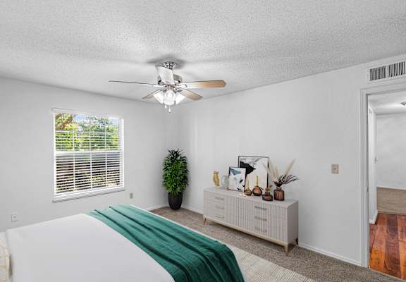 A bedroom with a bed, a window with blinds, a ceiling fan, and a side table with decorative items.