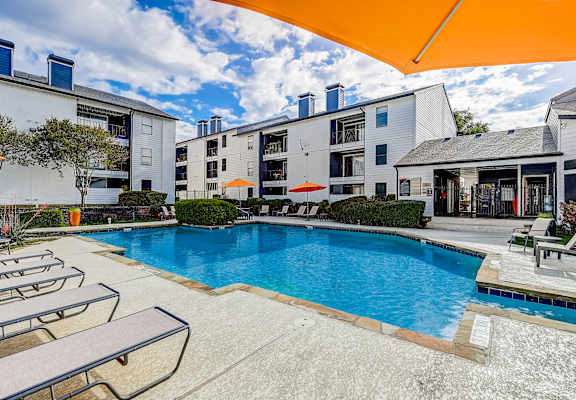 A swimming pool surrounded by sun loungers and umbrellas in front of apartment buildings.