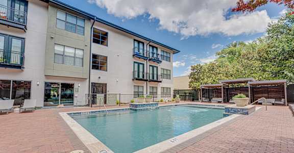 A swimming pool surrounded by a brick patio in front of apartment buildings.