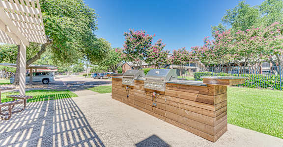A wooden structure with a grill on top is situated in a sunny outdoor area with trees and a car in the background.