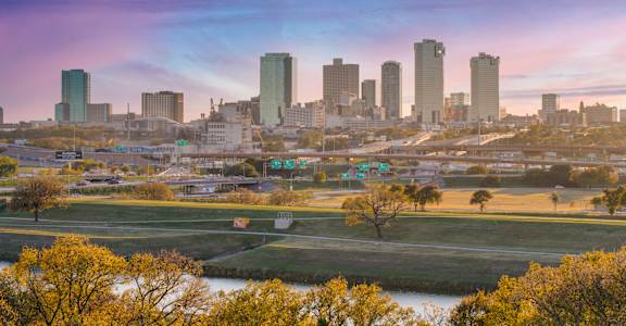 A city skyline with a river in the foreground and autumn trees.